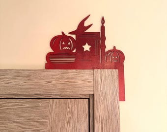 May include: A red wooden Halloween decoration featuring a witch's hat, a star, a candle, a stack of books, and two carved pumpkins. The decoration is placed on top of a wooden cabinet, against a pale yellow wall.