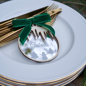 May include: A white plate with a gold rim, topped with a smaller white plate with a gold rim. A wooden Christmas ornament with a mirror finish is placed on the plates. The ornament is shaped like a Christmas tree and has the name "Gabriels" written on it. A green velvet ribbon is tied around the ornament.