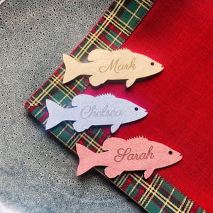 Könnte beinhalten: Drei fischförmige Holzornamente in hellem Holz, Hellgrau und Hellrosa. Jedes Ornament ist mit einem lasergravierten Namen in Schreibschrift versehen: Mark, Chelsea und Sarah. Die Fische haben ein kleines kreisförmiges Auge und sind diagonal angeordnet.