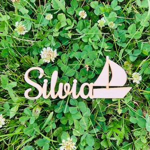 May include: A wooden cutout of the name "Silvia" with a sailboat design. The cutout is placed on a bed of green clover.