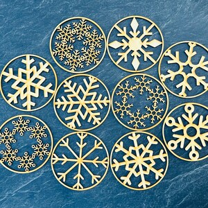 Peut inclure: Dix ornements en bois en forme de flocons de neige, chacun avec un design différent. Les ornements sont peints dans une couleur bois clair et sont disposés en cercle sur un fond bleu.