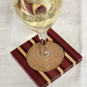 May include: A glass of white wine on a wooden coaster with a cork insert. The coaster is made of multiple wood types, including purpleheart, maple, and walnut.