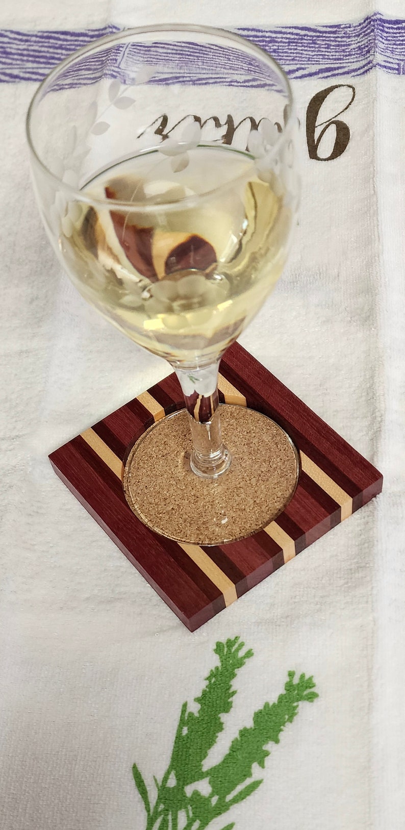 May include: A glass of white wine on a wooden coaster with a cork insert. The coaster is made of multiple wood types, including purpleheart, maple, and walnut.
