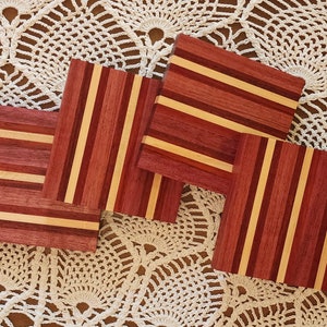 May include: Four square wooden coasters with a striped pattern in shades of red, brown, and beige. The coasters are arranged on a white crocheted surface.