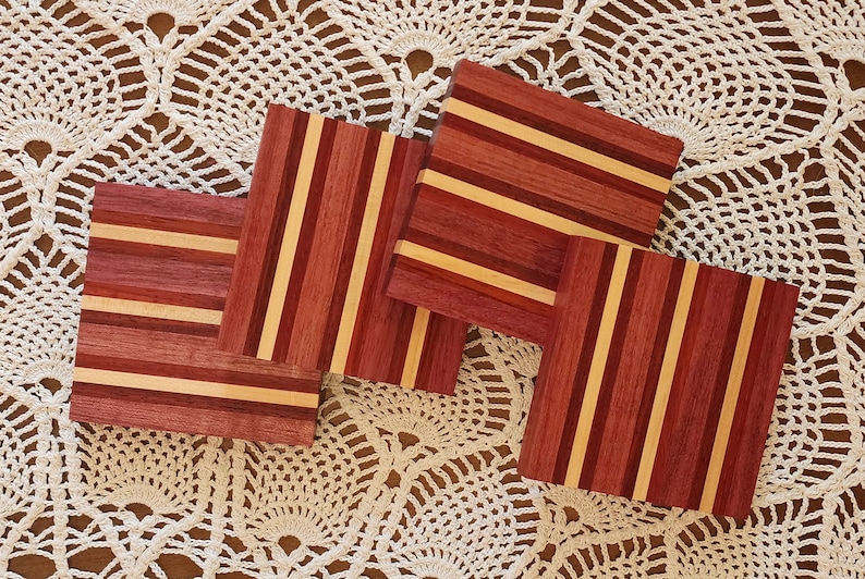 May include: Four square wooden coasters with a striped pattern in shades of red, brown, and beige. The coasters are arranged on a white crocheted surface.