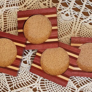 May include: Four wooden coasters with cork inserts. Each coaster has a square frame with alternating stripes of red, light brown, and dark brown wood.