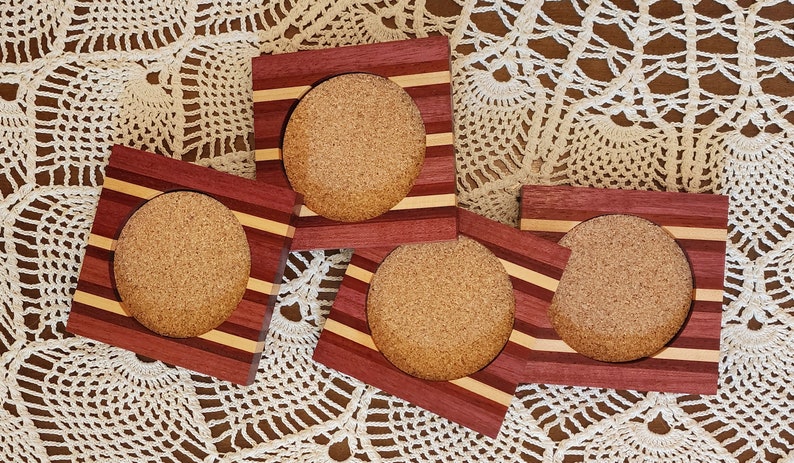 May include: Four wooden coasters with cork inserts. Each coaster has a square frame with alternating stripes of red, light brown, and dark brown wood.