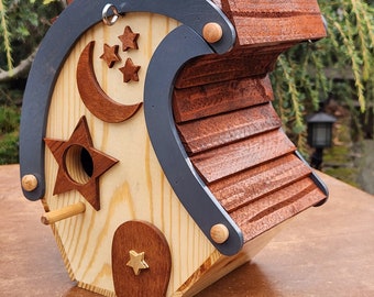 May include: Wooden birdhouse with a whimsical design. Features a curved roof, a crescent moon, stars, and a star-shaped entrance. The birdhouse is painted in natural wood tones, with accents of dark gray and brown.