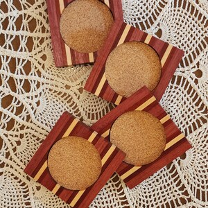 May include: Four square wooden coasters with cork inserts. The coasters have a red, brown, and light wood striped pattern.