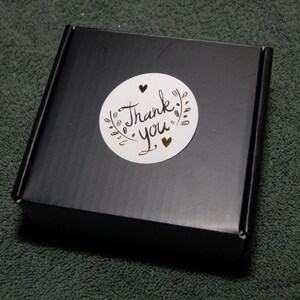 May include: A black box with a white circle sticker that says "Thank you" in gold lettering with two gold hearts.