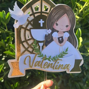 Pode incluir: Topper de bolo decorativo com uma menina de desenho animado em um vestido branco, uma pomba e um cálice. O topper tem detalhes dourados e o nome "Valentina" em letras douradas. O fundo é verde desfocado.