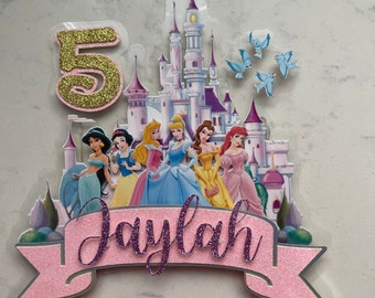 May include: A pink and gold cake topper featuring a castle, a banner with the name "Jaylah", and a number "5". The castle is surrounded by seven Disney princesses, including Cinderella, Belle, and Ariel. There are also blue birds flying around the castle.