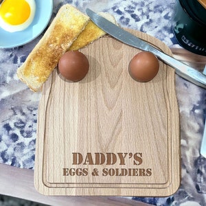 May include: A wooden serving board with two indentations for eggs and a slot for toast. The board is engraved with the text "Daddy's Eggs & Soldiers".