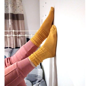 May include: Mustard yellow socks with ribbed detailing are worn with pink joggers. The socks are pulled up, creating a scrunched effect. The person is leaning against a white wall.