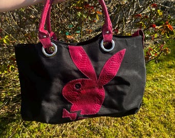 May include: Black tote bag with magenta accents and a magenta bunny logo. The bag has magenta handles and trim, with silver metal rings. The bunny logo is made of a textured material. The bag is held up against a background of green foliage.