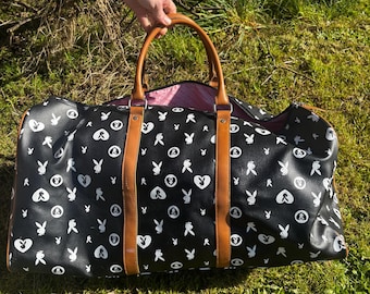 May include: Black duffel bag with white Playboy bunny and heart logos. The bag has brown leather handles and trim. The interior is pink. The bag is sitting on green grass.