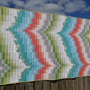 May include: A colourful quilt with a geometric pattern in shades of green, blue, red, and grey. The quilt is hanging on a wooden fence.