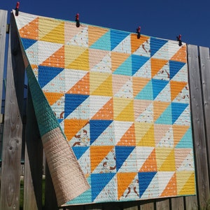 Op de afbeelding: Een quilt met een geometrisch patroon van oranje, gele, blauwe en witte driehoeken. De quilt hangt aan een waslijn buiten.