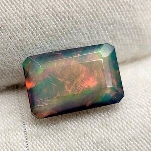 Puede incluir: Una piedra preciosa rectangular e iridiscente con un color base oscuro y destellos de rojo, naranja y verde. La gema está facetada con una superficie pulida, mostrando su juego de color interno. La piedra se sostiene sobre un fondo de tela blanca.