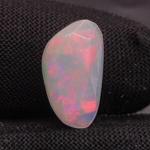 Peut inclure: Une pierre précieuse d'opale polie, de forme ovale, avec une surface facettée. L'opale présente un jeu de couleurs, avec des éclairs de rouge et de rose dans une base blanche translucide. La pierre est posée sur un fond sombre.