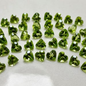 Może przedstawiać: Kolekcja zielonych peridotów w kształcie serca. Kamienie są fasetowane i mają jasny, żywy zielony kolor.
