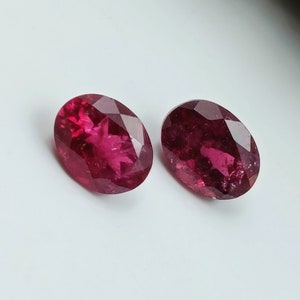 May include: Two oval-shaped, faceted gemstones with a deep pink hue. The gemstones are set against a white background.