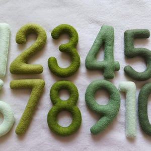 May include: Ten felt numbers in shades of green and white. The numbers are arranged in a row, starting with the number one and ending with the number ten.