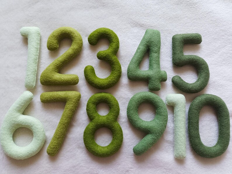 May include: Ten felt numbers in shades of green and white. The numbers are arranged in a row, starting with the number one and ending with the number ten.