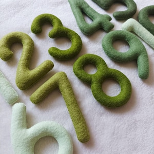 May include: A set of felt numbers in shades of green, from light to dark, with the number 6 in a lighter shade of green. The numbers are arranged in a random order on a white background.