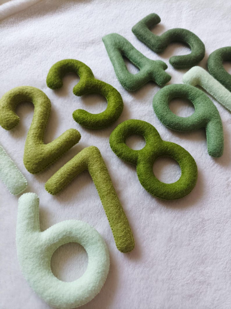May include: A set of felt numbers in shades of green, from light to dark, with the number 6 in a lighter shade of green. The numbers are arranged in a random order on a white background.