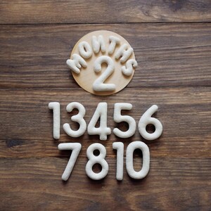 May include: White felt numbers on a wooden background. The numbers are 1, 3, 4, 5, 6, 7, 8, 10. A separate circle with the words "Months 2" is also on the background.