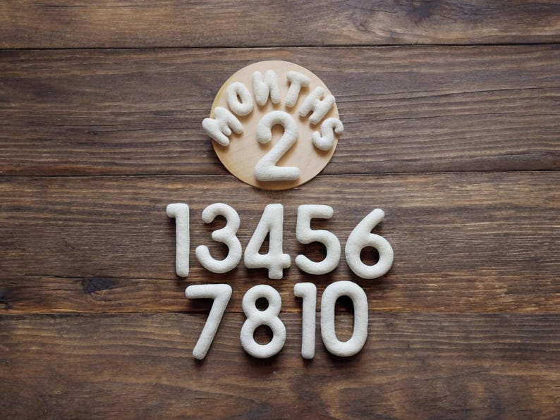 May include: White felt numbers on a wooden background. The numbers are 1, 3, 4, 5, 6, 7, 8, 10. A separate circle with the words "Months 2" is also on the background.