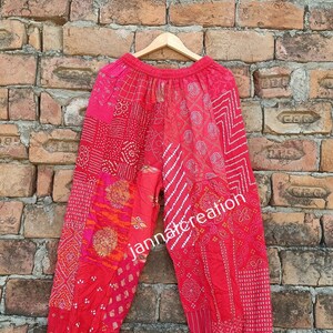 Może przedstawiać: Czerwone spodnie w patchwork z sznurkiem w pasie i elastycznymi mankietami. Spodnie są wykonane z różnych tkanin o różnych wzorach, w tym grochy, paski i wzory kwiatowe. Spodnie wiszą na drewnianym wieszaku na tle ceglanej ściany. Na spodniach widoczny jest tekst "jannatcreation".