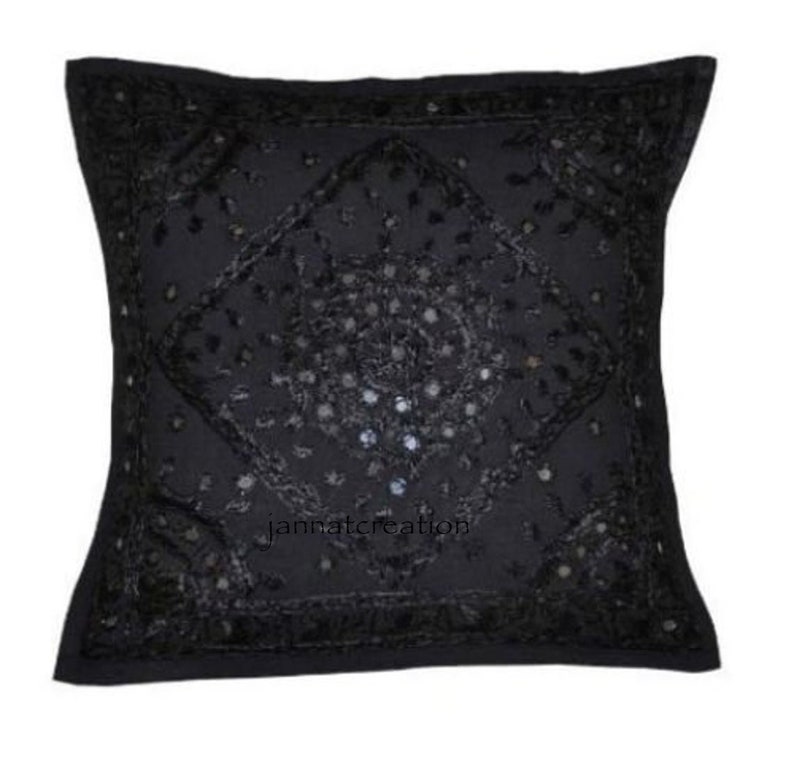 Puede incluir: Una funda de coj&iacute;n cuadrada de color negro con intrincados bordados negros y peque&ntilde;os espejos reflectantes dispuestos en un patr&oacute;n de diamante central. La superficie presenta costuras detalladas y el texto jannatcreation.