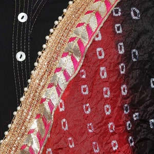 May include: Close-up of a black garment with white buttons and stitching. A decorative border features gold, pink, and white accents, with a red and black patterned fabric on the right side. The fabric has a square pattern.