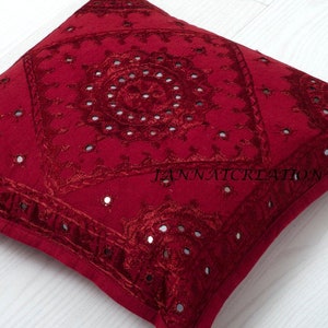 Puede incluir: Una funda de coj&iacute;n cuadrada de color rojo intenso con intrincados bordados tonales y peque&ntilde;os espejos circulares incrustados en la tela. El dise&ntilde;o incluye un patr&oacute;n central tipo mandala y el texto JANNATCREATION impreso en negro.