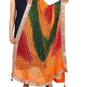 May include: A colorful dupatta with a bandhani pattern in shades of red, green, and orange. The scarf has a gold border with intricate detailing and small tassels. The fabric is draped, showcasing the vibrant colors and design.