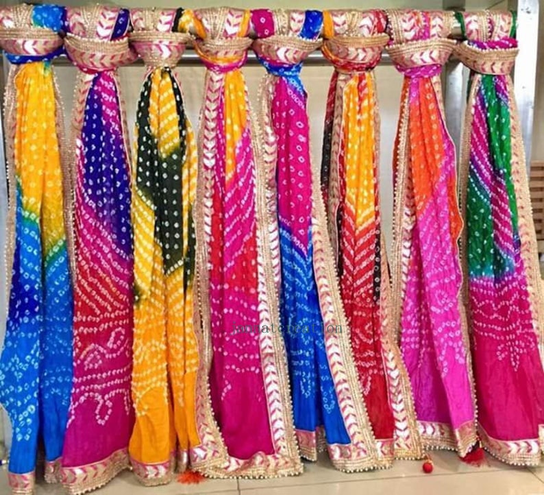 May include: A display of colorful, patterned scarves. The scarves are in shades of yellow, blue, purple, pink, orange, and green. Each scarf has a decorative gold trim. The scarves are hanging from a metal rod.