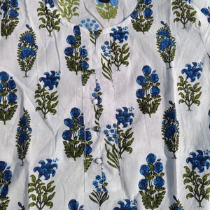 Puede incluir: T&uacute;nica de algod&oacute;n blanco con estampado floral azul y verde. La t&uacute;nica tiene un escote redondo y un cierre de bot&oacute;n.