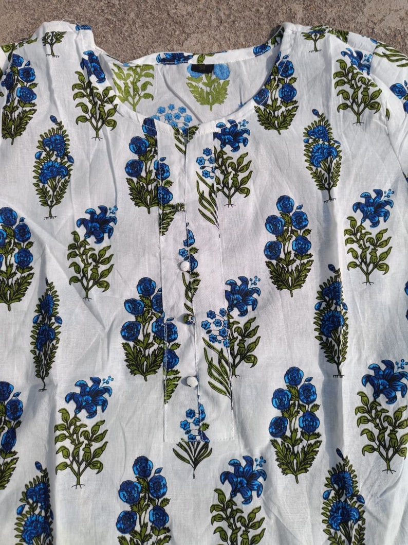 Puede incluir: T&uacute;nica de algod&oacute;n blanco con estampado floral azul y verde. La t&uacute;nica tiene un escote redondo y un cierre de bot&oacute;n.