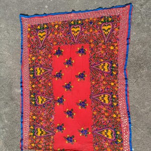 May include: A vibrant red textile with intricate embroidery. The design features heart-shaped motifs, floral patterns, and a blue border. The embroidery incorporates yellow, green, and blue threads, creating a colorful and detailed piece.