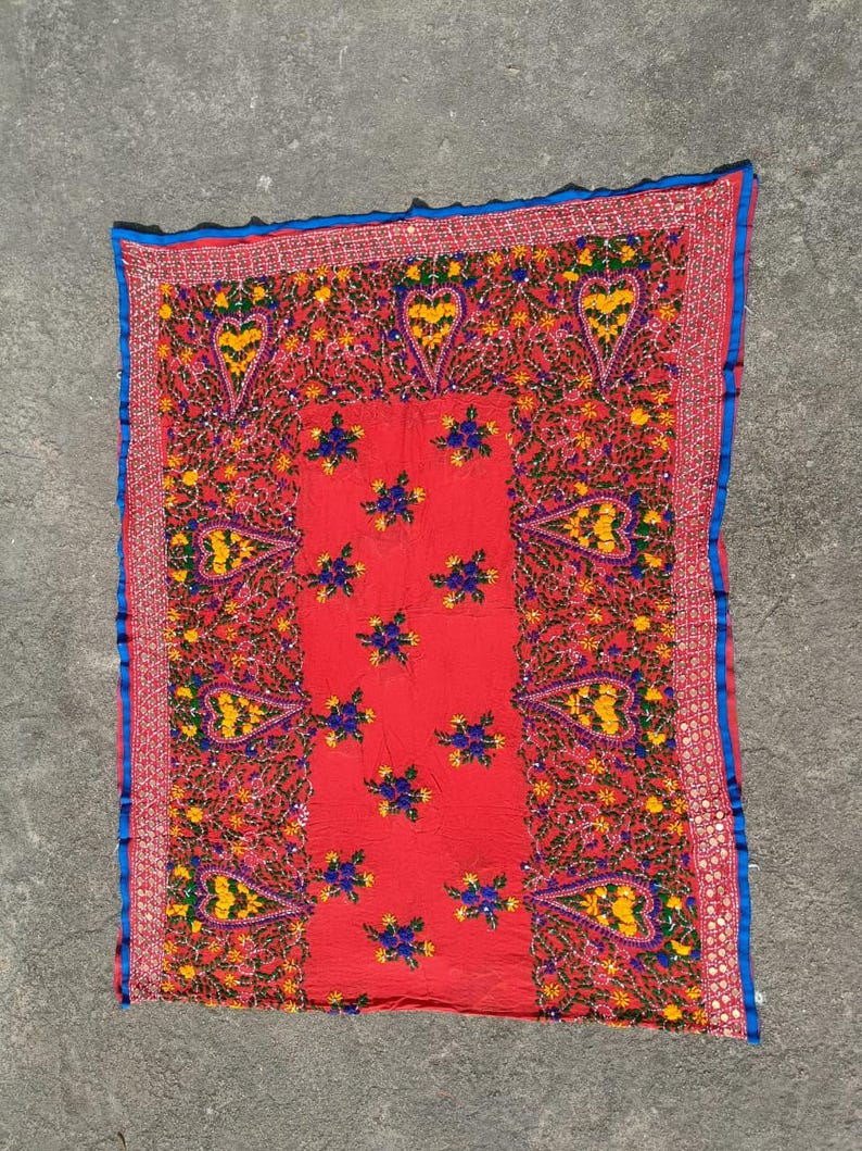 May include: A vibrant red textile with intricate embroidery. The design features heart-shaped motifs, floral patterns, and a blue border. The embroidery incorporates yellow, green, and blue threads, creating a colorful and detailed piece.