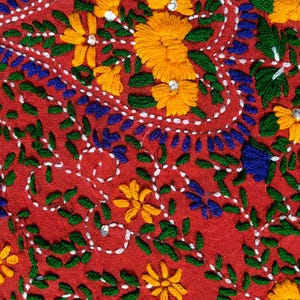 May include: Close-up of red fabric with intricate embroidery. The design features yellow flowers, blue and green leaves, and white dotted lines. Small, shiny embellishments are scattered throughout the floral pattern.