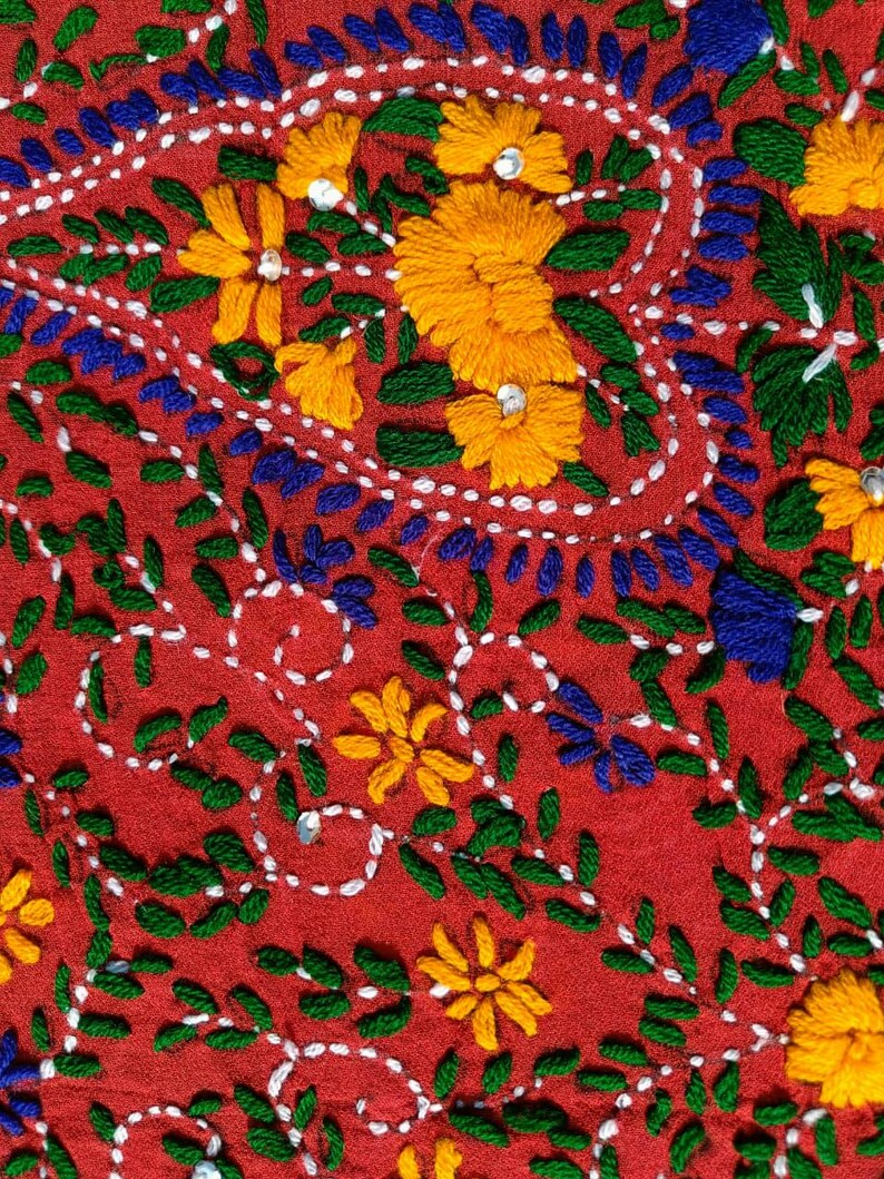 May include: Close-up of red fabric with intricate embroidery. The design features yellow flowers, blue and green leaves, and white dotted lines. Small, shiny embellishments are scattered throughout the floral pattern.