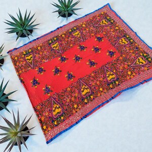 May include: A vibrant red textile with intricate embroidery. The square fabric features a central panel with floral and paisley designs in yellow, blue, and orange. A blue border frames the edges. The textile is likely a decorative item or scarf.