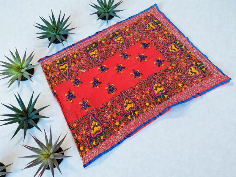 May include: A vibrant red textile with intricate embroidery. The square fabric features a central panel with floral and paisley designs in yellow, blue, and orange. A blue border frames the edges. The textile is likely a decorative item or scarf.