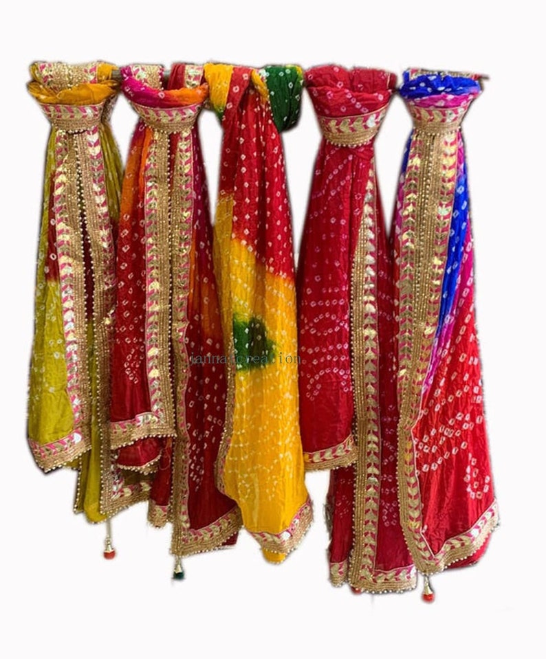 May include: A collection of colorful, patterned scarves hanging against a white background. The scarves feature a variety of colors, including yellow, red, green, and blue, with gold trim and tassels. The fabric has a dotted pattern.