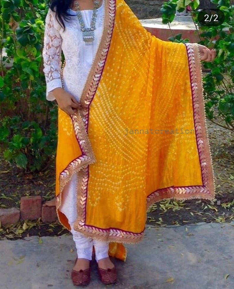 May include: A woman's white embroidered tunic and leggings paired with a vibrant yellow dupatta. The dupatta features a detailed pattern and a decorative border. The text "jannatcreation" is visible on the dupatta.