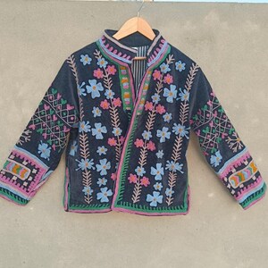May include: A dark charcoal jacket featuring vibrant, multi-colored floral embroidery across the front and sleeves. The garment has a high collar, long sleeves, and a black and white striped interior lining, all displayed on a wooden hanger.