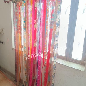 Può includere: Una tenda colorata composta da strisce di tessuto verticali in vari modelli e tonalità di rosa, rosso, arancione e blu. La tenda è appesa davanti a una finestra, con il testo "jannatcreation" visibile sul tessuto.